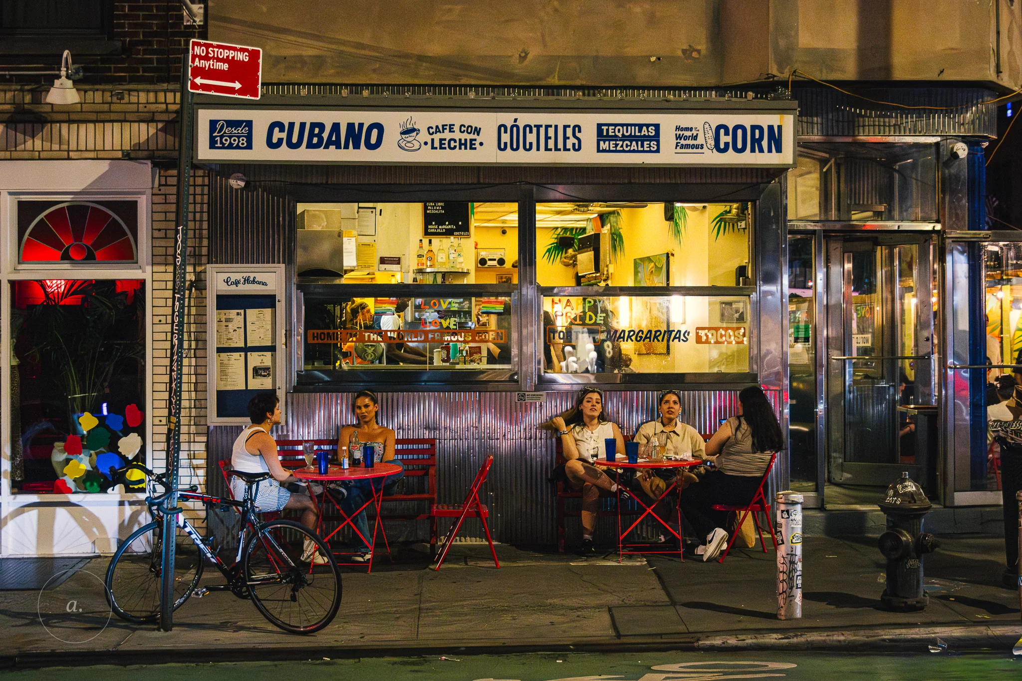 The lit front of a late night Cuban takeout window, its CUBANO sign glowing above diners at sidewalk tables, photographed by Campbell Jensen